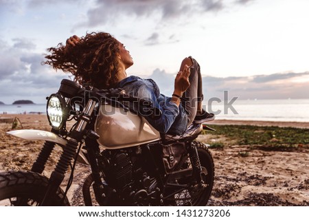Beautiful girl having fun driving her custom cafe racer motorcycle, enjoying the sunset on the beach