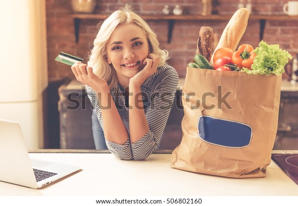 Beautiful girl is holding a credit card, leaning\
on table near paper shopping bag with food and a laptop, looking at\
camera and smiling