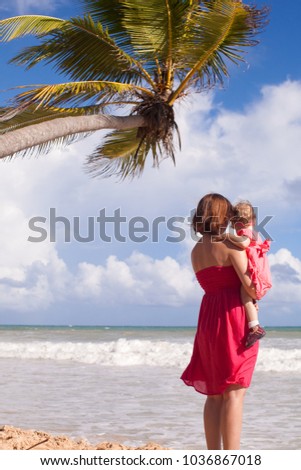 a beautiful girl is holding a little girl in her arms and looking out into the distance to the sea and clouds, and above them is an exotic palm tree