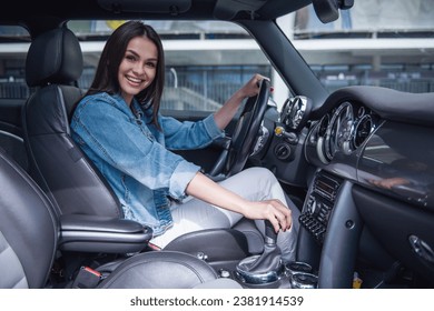 Beautiful girl in jean jacket is looking at camera and smiling while sitting in her car