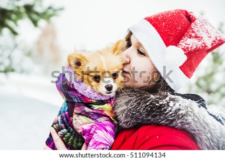 Beautiful girl kissing dogs. The woman with the German Spitz. Fairy tale girl embracing cute dog in winter park. Christmas. Female wear red cap of Santa Claus 