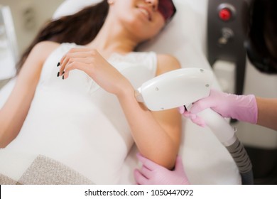 A beautiful girl lies in the office of a beautician in red goggles from laser irradiation, the hands of the doctor remove hair on hands with a laser device