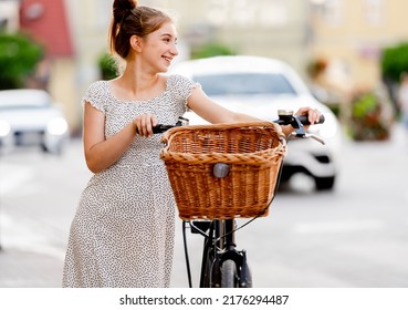 Beautiful Girl In Long Dress Walking At Street In City With Bicycle And Looking Back. Pretty Female Teenager Model Outdoors In Summer Time With Basket Bike