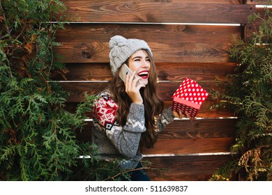 Beautiful Girl With Long Hair With Christmas Box On Wooden Background Surround Green Branches. She Wears Warm Winter Clothes, Speaking On Phone, Laughing To Side