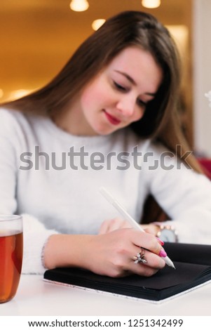 a beautiful girl with long hair sitting in a cafe and writing something in a notebook with black pages