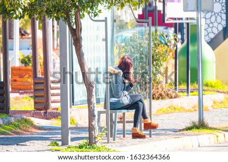 Beautiful girl with long hair is sitting on the bus stop bench with a smart phone in his hand