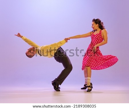 Beautiful girl and man in colorful retro style costumes dancing incendiary dances isolated on lilac color background in neon light. Actors in motion and action. Concept of art, 60s, 70s culture