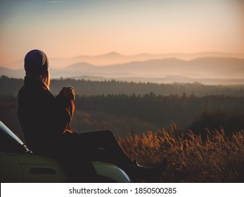 Beautiful girl with mug of tea sitting on a car in beautiful mountains Beskides in November in sunset - Shutterstock ID 1850500285