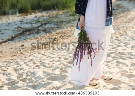 Beautiful girl on the beach. Dresses in boho style. In the hands of a woman a bouquet of wild flowers. Wind, Sunset, coast. copy space. sand