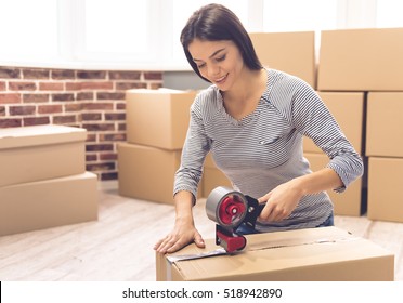 Beautiful girl is packing a moving box using an adhesive tape looking at camera and smiling