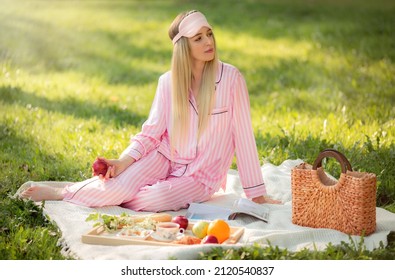 Beautiful girl in pink pajamas is having breakfast in nature. She is sitting on the grass on a blanket, next to her a tray with delicious food and tea. She eats an apple and reads a magazine. 