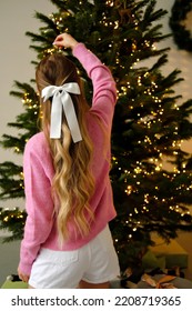 A Beautiful Girl In A Pink Sweater, Shorts, White Leggings And A Hair Clip In The Form Of A Bow In A Cozy Home Interior Decorates The Christmas Tree. 
 Vertical Photo