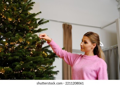 A Beautiful Girl In A Pink Sweater, Shorts, White Leggings And A Hair Clip In The Form Of A Bow In A Cozy Home Interior Decorates The Christmas Tree. Horizontal Photo