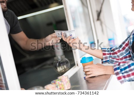 A beautiful girl in a plaid shirt buys coffee in a diner. The seller gives her coffee, she gives the seller money for coffee