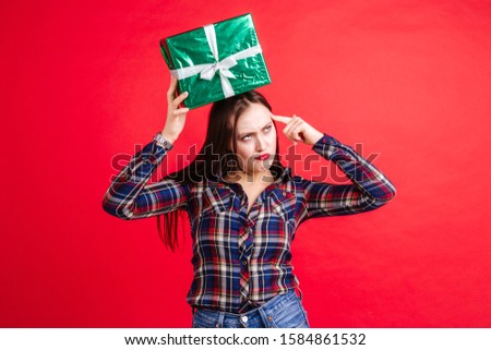 Beautiful girl in a plaid shirt holding a gift box on her head standing on a red background holding a finger to her temple showing that fool