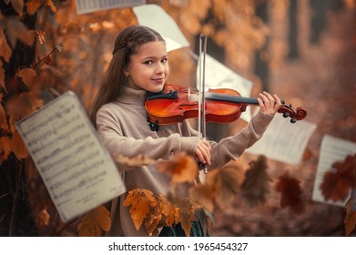 A beautiful girl plays the violin among the autumn orange foliage in the forest and with music sheets in the background.