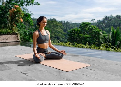 A beautiful girl practices yoga by the pool in the morning in Bali, Indonesia. Young woman in sportswear near the infinity pool against the backdrop of a tropical landscape.