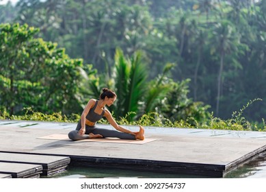 A beautiful girl practices yoga by the pool in the morning in Bali, Indonesia. Young woman in sportswear near the infinity pool against the backdrop of a tropical landscape.