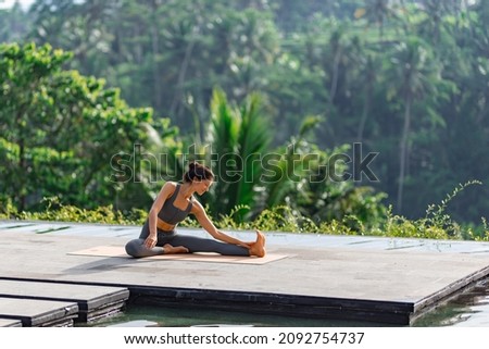 
A beautiful girl practices yoga by the pool in the morning in Bali, Indonesia. Young woman in sportswear near the infinity pool against the backdrop of a tropical landscape.