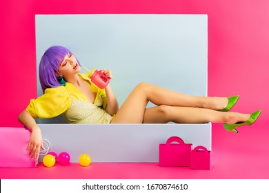 beautiful girl in purple wig as doll drinking from jar while sitting in blue box with balls and shopping bags, on pink