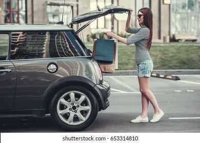 Beautiful Girl Is Putting Shopping Bags Into Car Trunk And Smiling