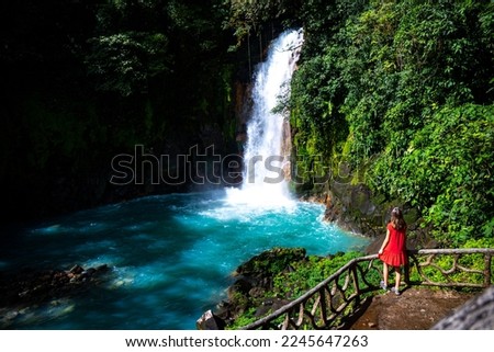 A BEAUTIFUL GIRL IN A RED DRESS STANDS UNDER THE FAMOUS BLUE WATERFALL OF RIO CELESTE, COSTA RICA; A WATERFALL WITH BLUE WATER IN VOLCANO TENORIO NATIONAL PARK; A WATERFALL IN THE RAINFOREST	