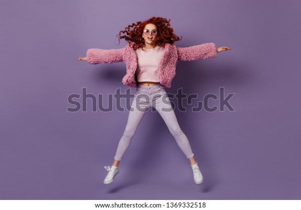 Beautiful girl with red hair is jumping against\
lilac background. Portrait of woman dressed in skinny white pants\
and pink jacket