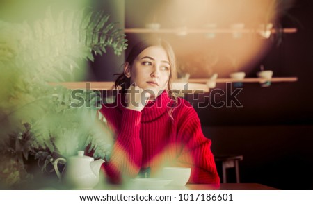 A beautiful girl in a red knitted sweater drinks tea and reads a book in a cozy cafe, the concept of pleasant leisure and communication