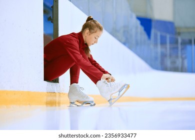 Beautiful girl in red sportswear preparing for training session on ice rink arena, figure skater tying laces on skates. Concept of professional sport, competition, sport school, health, hobby, ad