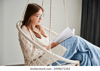 Beautiful girl relaxing and reading book on indoor swing. Pretty female in glasses excited about story she reading. Cozy afternoon with book in hanging chair in home. - Powered by Shutterstock