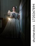 Beautiful girl, in a romantic dress, retro style.With dark hair. Stands tall with a fan against the background of the stage in the theater, and looks away. Historical reconstruction                   