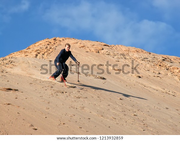 Beautiful\
girl Sand skiing down dunes in desert. Sand-skiing is sport and\
form of skiing in which skier rides down sand dune on skis, using\
ski poles, as done with other types of\
skiing.