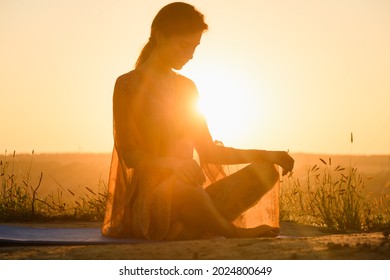 Beautiful girl sit in yoga lotus pose against sun in warm light, wear transparent cloth, meditation and stretching outdoors