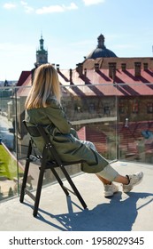 A beautiful girl sits on the balcony and enjoys the city view on a sunny day. Calm lifestyle. Happy woman resting after work on the outdoor terrace. Nice view of the city from the balcony. Back view