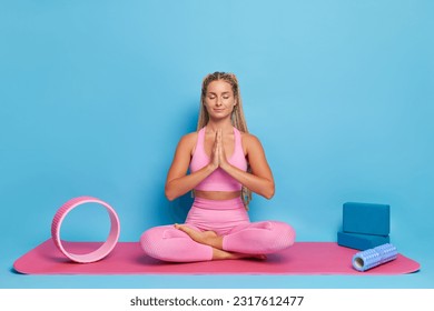 Beautiful girl sits on pink mat in yoga lotus pose dressed in pink top and leggings, sports equipment lies nearby, fitness concept, copy space - Powered by Shutterstock