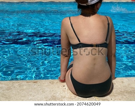 A beautiful girl sits in a swimsuit at the edge of the pool, view from the back. Concept: vacation in a tropical resort in a hotel.