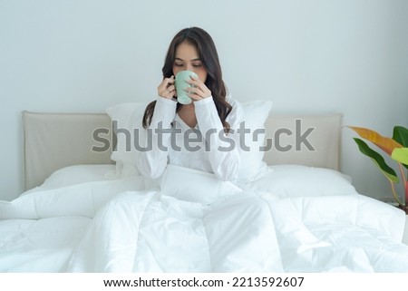 Beautiful girl sitting and drinking hot tea in the morning On a soft white bed Feel relaxed under the warm blankets.