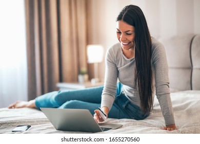 Beautiful girl sitting on the bed surfing the web on her laptop and using credit card for online shopping.
