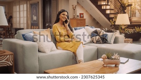 Beautiful Girl Sitting On A Couch At Home, Talking on a Phone, Chatting With Her Family, Friends, Girfriends Or Boyfriend. Happy Sweet Indian Lady Uses Mobile Phone For Communication with Loved Ones