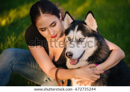 A beautiful girl is sitting on the lawn with her dog. Siberian husky dog with blue eyes. Bright green trees and grass are in the background. Friendship between man and animals.