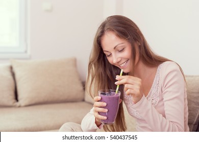 Beautiful girl sitting on a living room couch, smiling and holding a glass of raspberry and blueberry mix smoothie - Powered by Shutterstock