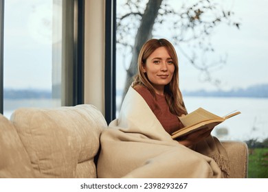 A beautiful girl, sitting on the sofa, reading a book and wrapped in a blanket.