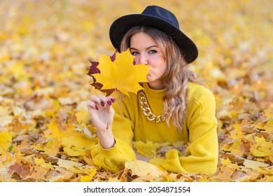 Beautiful Girl  Smile, Young Happy Woman  In Black Hat Lying In Maple Leaves In Autumn In The Park. Portrait Of Stylish Smiling Lady In Yellow Clothes. Fashion Female Fall Outdoors. Copy Space, Text