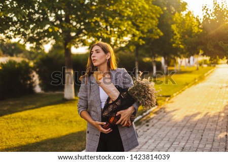 Beautiful girl in square jacket hold of flowers, string bag with fruits and baguette go to home. Romantic girl come to private house with trees. Lifestyle concept. Woman go to home at evening.