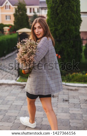 Beautiful girl in square jacket hold of flowers, string bag with fruits and baguette go to home. Romantic girl come to private house with trees. Photo from back and girl is turn around. Housewife.