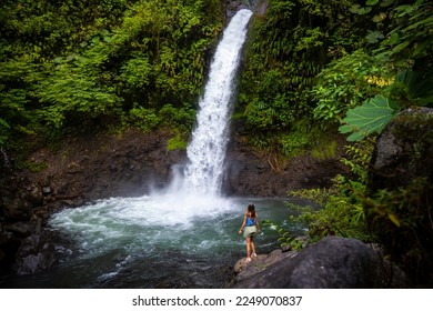 A beautiful girl stands on rocks under a powerful tropical waterfall in Costa Rica; la paz waterfall in the jungle	

