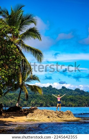 A beautiful girl stands on the rocks under a palm tree on the shore of a Costa Rican beach in the Caribbean; vacation on a paradise beach in Costa Rica