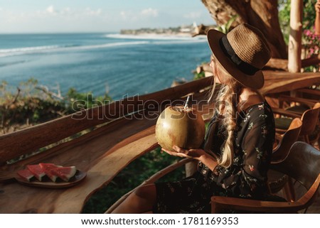 Beautiful girl in straw hat with coconut cocktail in beach cafe with ocean view. Tropical vacation concept