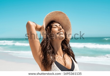 Beautiful girl with straw hat enjoying sunbath at beach. Close up face of young tanned woman with closed eyes enjoying breeze at seaside. Carefree latin woman smiling with ocean in background.