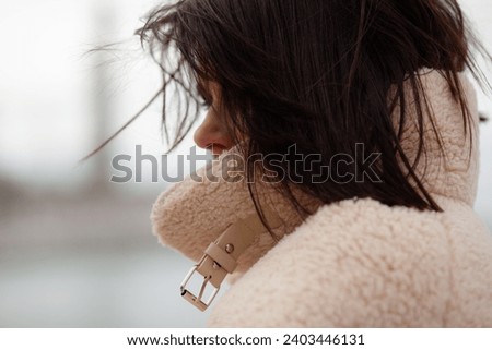 A beautiful girl with a stylish bob haircut is walking around the city, against the background of water. The woman is wearing a white faux fur sheepskin coat 